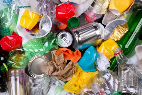 A close-up view of a large pile of mixed recyclable waste plastic bottles, aluminium cans, and crumpled paper, all discarded haphazardly on a surface. The plastic bottles vary in color, including green, clear, and colored containers, some with caps still attached. The aluminium cans are predominantly silver, with some crushed and others intact, positioned among the plastic bottles and paper. Crumpled brown paper and brightly colored plastic wrappers are also visible within the heap, creating a chaotic mix of materials. The waste is spread across a concrete or pavement surface, suggesting an outdoor setting typical of rubbish collection or disposal areas in South East London, such as Dulwich, where Commercial Waste Dulwich offers business waste removal services. The lighting appears natural, emphasizing the reflective surfaces of the cans and the translucent quality of the plastic bottles, highlighting the variety of waste materials that an effective rubbish removal service would manage among commercial or residential environments in the area.